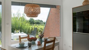 A dining area with a table set for two, a wicker pendant light, and a view of a brick wall and greenery outside.