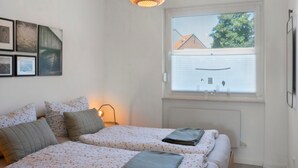 A bedroom with two beds, a window with blinds, a wall-mounted lamp, and framed pictures.