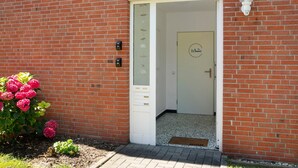 A brick building with a white door, a small garden with pink flowers, and a light fixture on the wall.