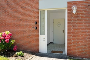 A brick building with a white door, a small garden with pink flowers, and a light fixture on the wall. - Apartment 'Weitblick - In 5 Min Am Meer' with Private Terrace, Private Garden and Wi-Fi (Wangerland)