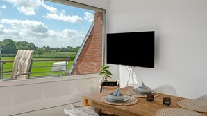 Ein Zimmer mit großem Fenster, einem an der Wand befestigten Flachbildfernseher, einem Holztisch mit Pflanze und Blick auf einen Ziegelkamin und eine grüne Landschaft.