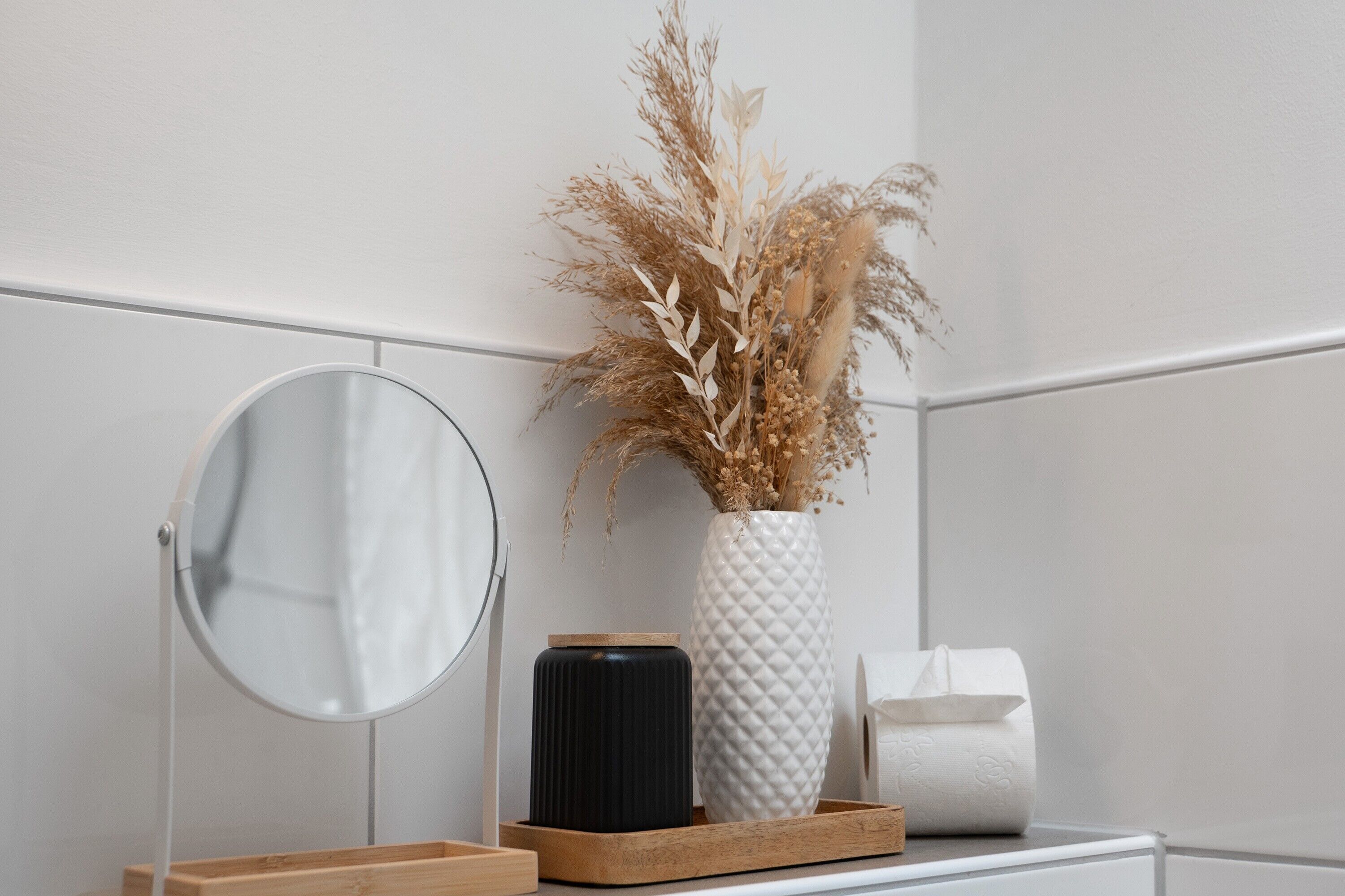 A bathroom with a round mirror, a white vase with dried plants, a black container, and a wooden tray.
