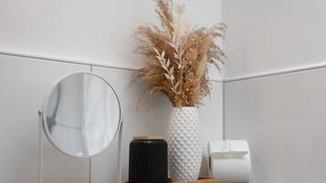A bathroom with a round mirror, a white vase with dried plants, a black container, and a wooden tray.