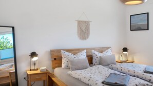 Ein Schlafzimmer mit einem hölzernen Kopfteil, zwei Nachttischen mit Lampen, einem Wandbehang aus Makramee und einem gerahmten Bild an der Wand.