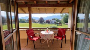A covered balcony with two red chairs and a table, overlooking a grassy area with houses and mountains. - Apartment '2' with Mountain View, Shared Garden and Wi-Fi (Krün)