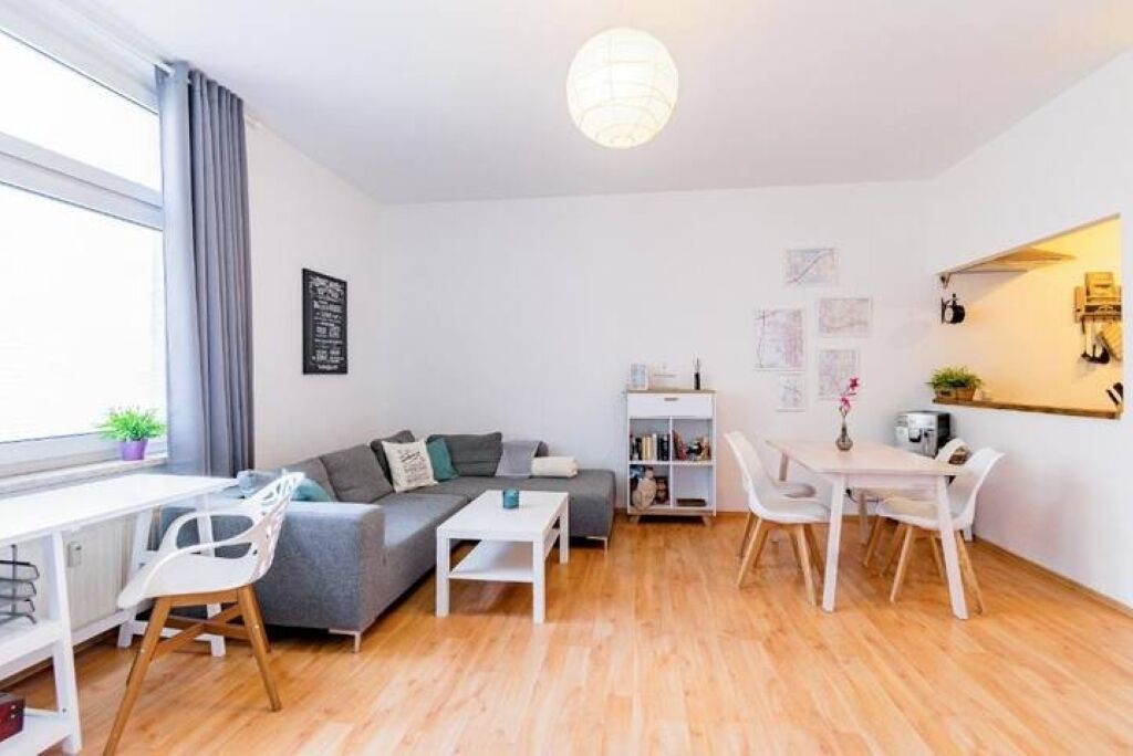 A modern living room with a sofa, dining table, and chairs. There is a bookshelf and a window with curtains.