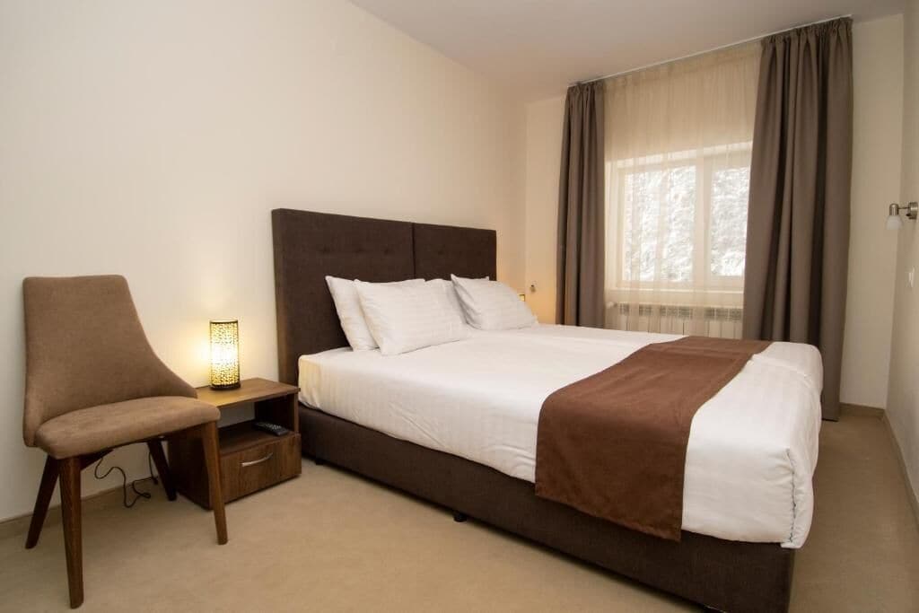 Una habitación de hotel con una cama, una silla, una mesita de noche con lámpara y un ventanal con cortinas.