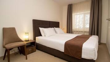 Una habitación de hotel con una cama, una silla, una mesita de noche con lámpara y un ventanal con cortinas.