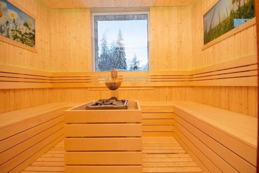 Una sauna con paredes y bancos de madera, una ventana con vistas a árboles, y un brasero central.