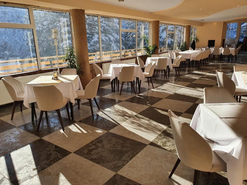 A spacious dining area with checkered flooring, white tables, and chairs, large windows with a snowy mountain view.