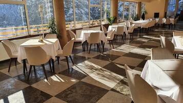 A spacious dining area with checkered flooring, white tables, and chairs, large windows with a snowy mountain view.