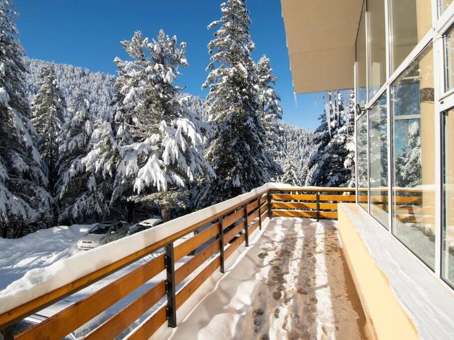 A balcony with a wooden railing overlooking a snowy landscape with trees and a clear blue sky.