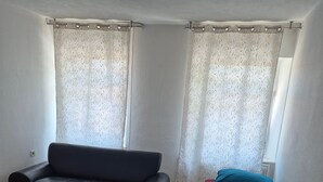 A room with a black sofa, a single bed with a red and blue pillow, and a window with patterned curtains. - Apartment 'Appartement Aurach A6' with Wi-Fi (Aurach)