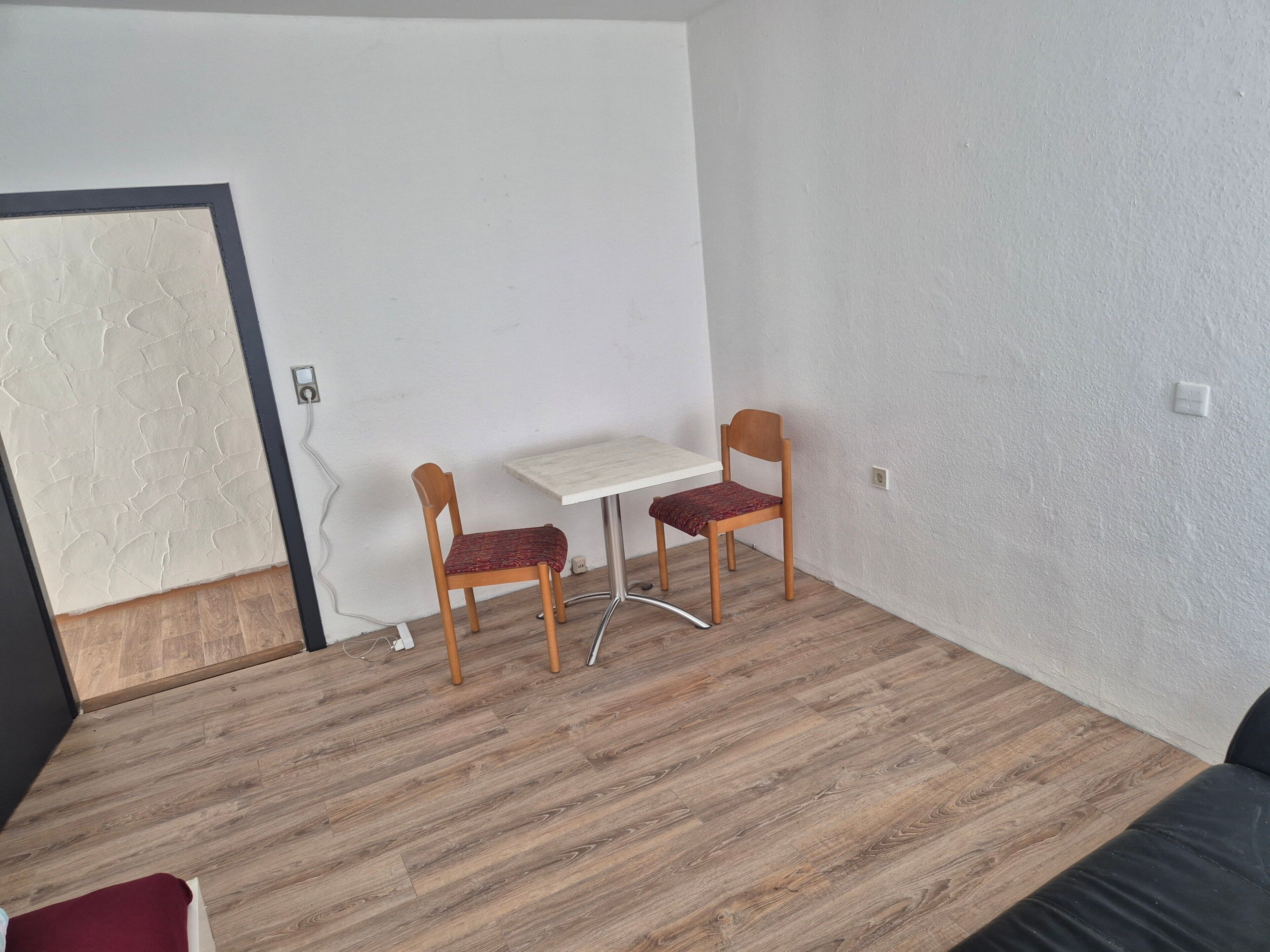 Una habitación con piso de madera, pared blanca, una pequeña mesa con una silla y una puerta con diseño texturizado.