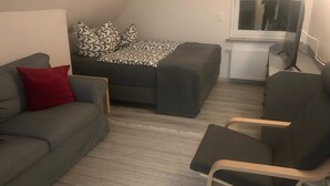 A room with a grey sofa, a red pillow, a patterned bed, a small table, and a chair.