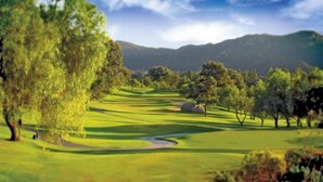 Campo de golf con un obstáculo acuático, árboles y colinas onduladas al fondo.