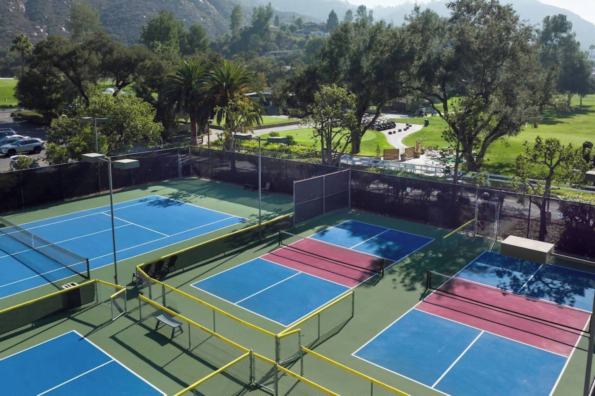A tennis court with multiple courts, surrounded by trees and a mountainous backdrop.
