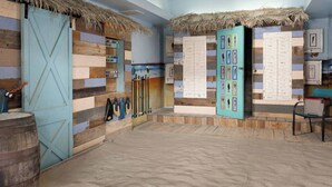 Habitación con pared de madera, techo de paja y suelo arenoso. Hay dos puertas, una con marco azul y otra con marco blanco.