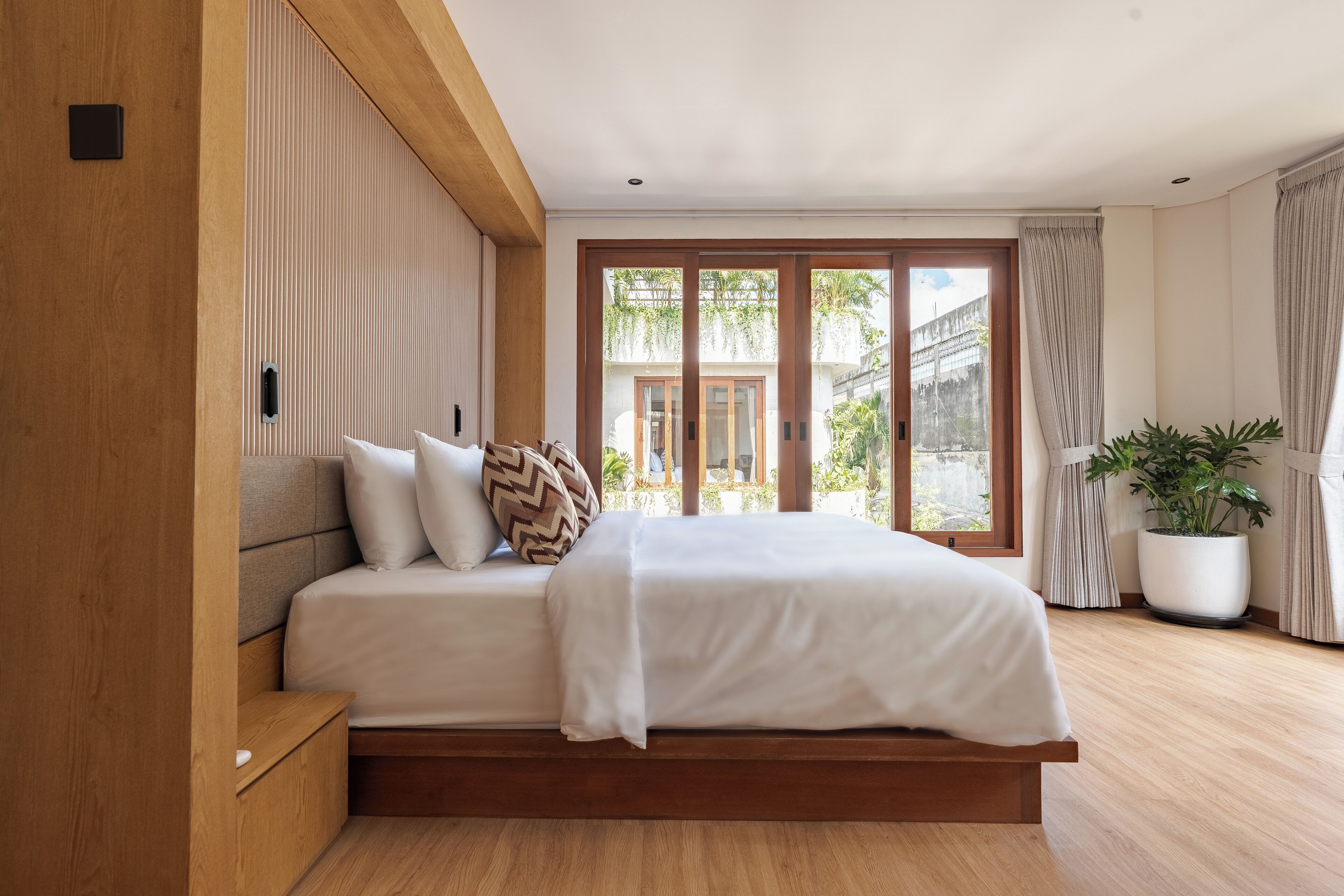 A modern bedroom with a large bed, wooden headboard, and a sliding glass door leading to a balcony.