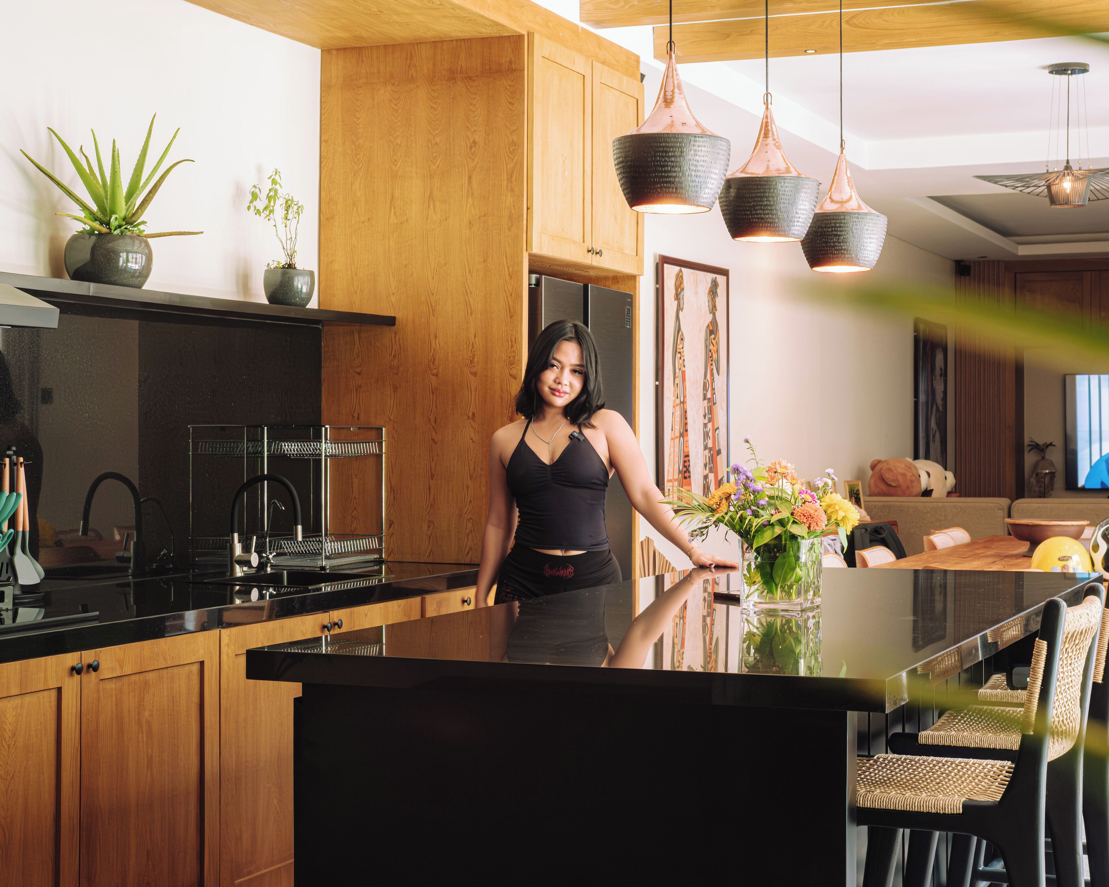A modern kitchen with wooden cabinets, a black countertop, and a central island. There are hanging pendant lights, a vase with flowers, and a dining area with wicker chairs.