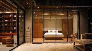 Une chambre moderne avec un grand lit, des meubles en bois et une porte en verre donnant sur un espace de vie doté d’étagères.