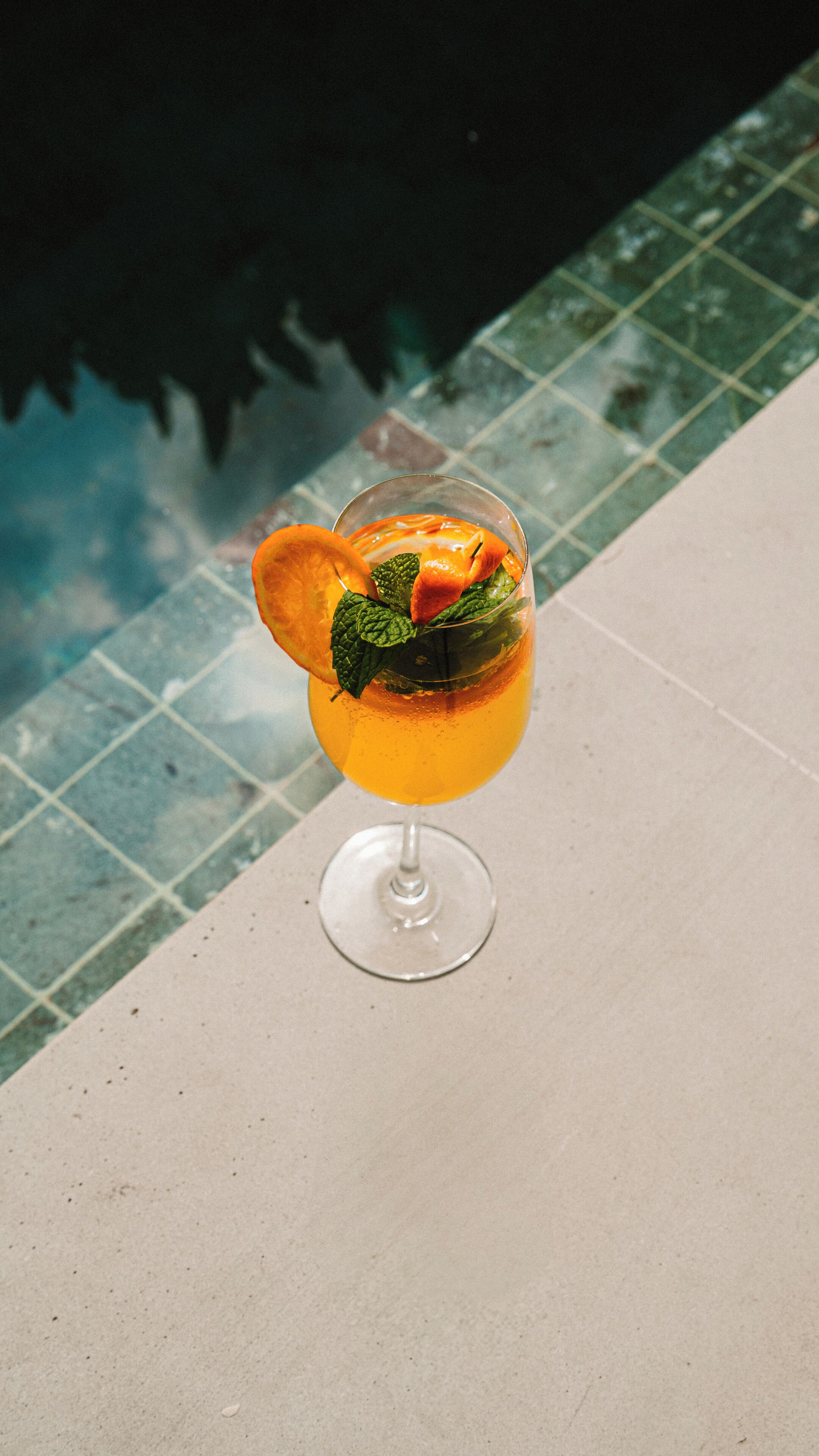 A cocktail with orange slices and mint leaves on a tiled poolside.