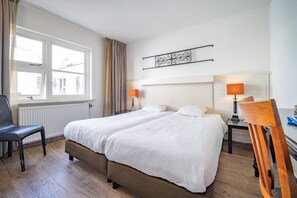 Two single beds with white linens, each with a bedside table and lamp, are positioned in this hotel room, accompanied by a desk and chair.