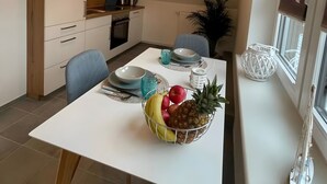 Un comedor moderno con una mesa blanca, sillas y un frutero. Se observa una cocina al fondo con armarios y una lámpara colgante.
