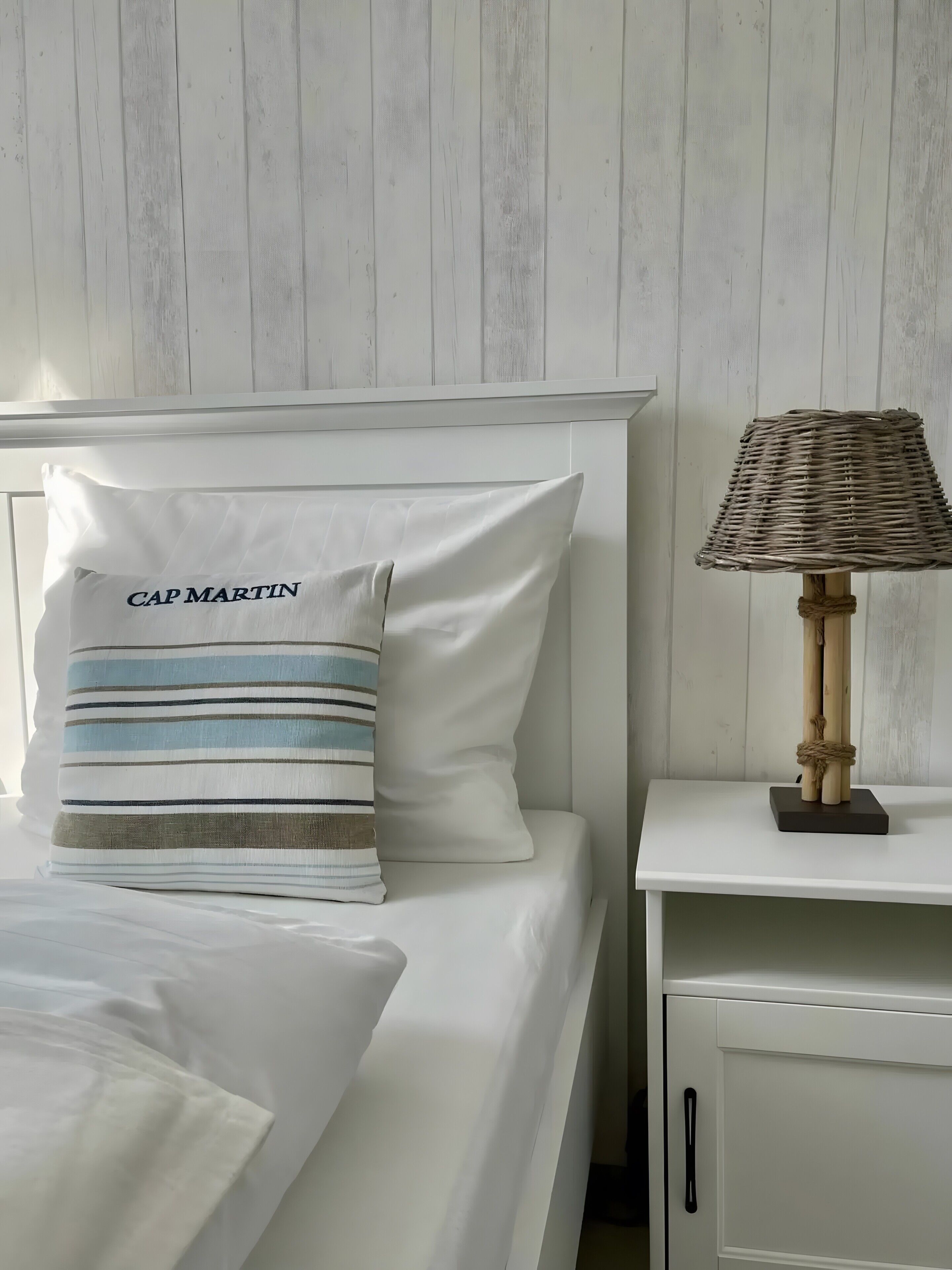 Ein Bett mit weißer Bettwäsche, ein Kopfkissen mit dem Aufdruck 'CAP MARTIN', ein gestreiftes Kissen und eine Korbtasche als Lampe auf einem Nachttisch.