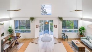 A spacious living area with a wooden floor, a dining table with chairs, a sofa, and two large windows offering a view of the outdoors. - 6 Bedroom Oasis in Burleigh Heads (Burleigh Heads)