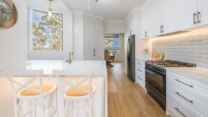 A modern kitchen with white cabinetry, a central island with bar stools, a built-in stove, and a large window with a view of trees. - 6 Bedroom Oasis in Burleigh Heads (Burleigh Heads)