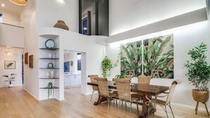 A spacious dining area with a wooden table and wicker chairs, surrounded by large tropical plant paintings. - 6 Bedroom Oasis in Burleigh Heads (Burleigh Heads)