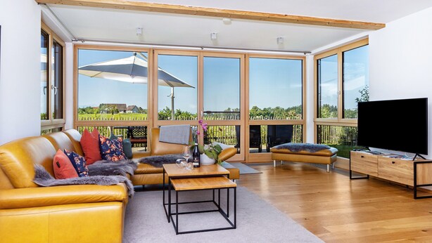 Ein geräumiges Wohnzimmer mit einem gelben Sofa, einem Flachbildfernseher und großen Fenstern mit Blick auf eine grüne Landschaft.