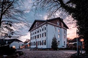 Traditional European-style building with a central entrance and gabled roof, set in a serene cobblestone courtyard surrounded by mature trees.
