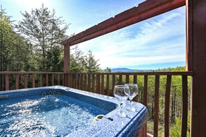 A hot tub with two glasses on a wooden deck overlooking a forest and mountains.
