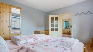 Un dormitorio con dos camas, un armario de madera, una puerta que da a otra habitación y una mesita con una silla.