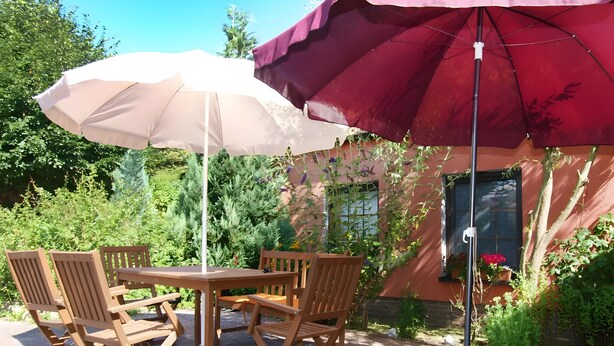 Un patio al aire libre con una mesa, sillas, un parasol grande y un edificio con ventana y plantas.