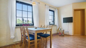 Una habitación con una mesa de comedor de madera, cuatro sillas, dos ventanas con cortinas, un televisor montado en la pared y una puerta.