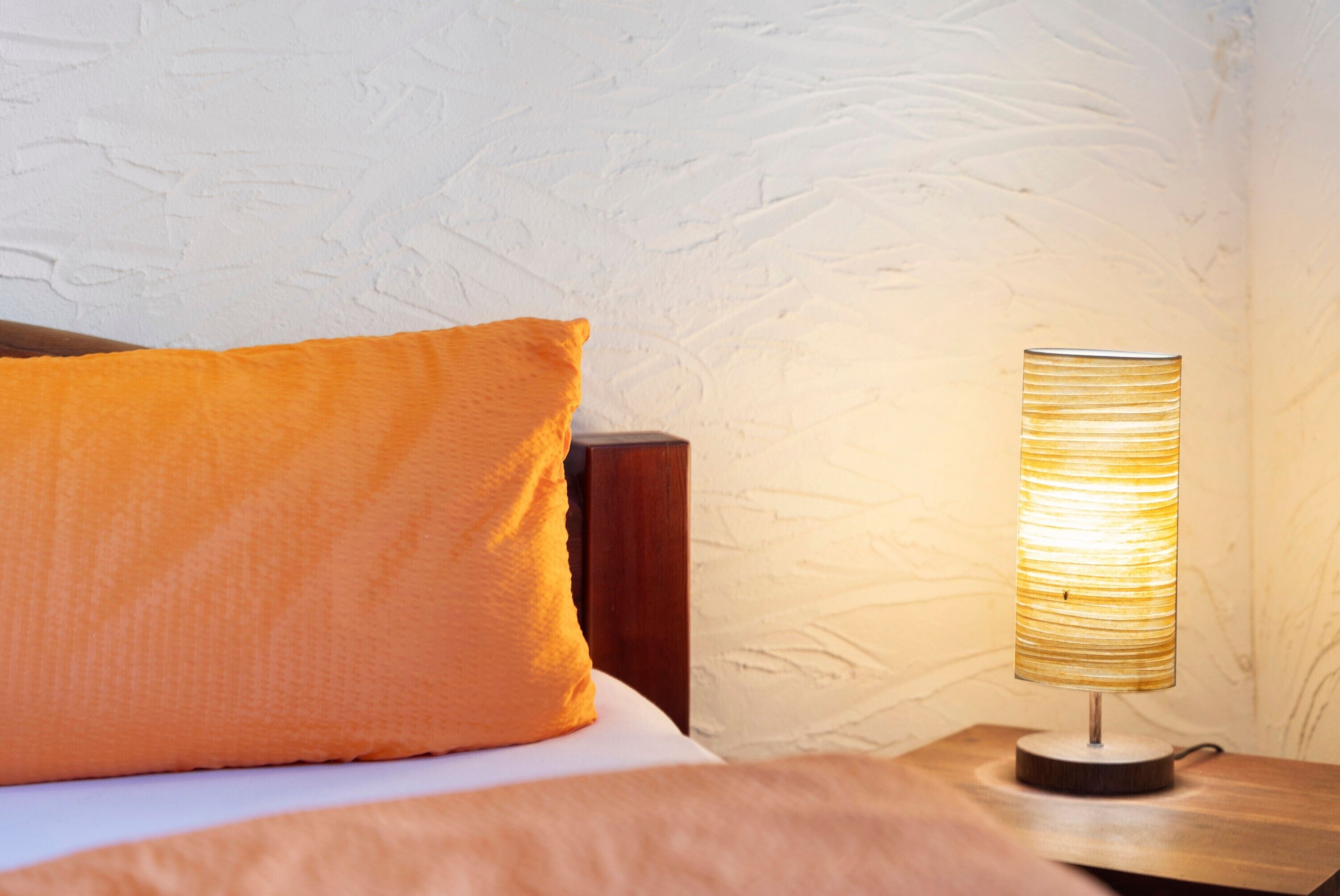 Un letto con un cuscino arancione e un comodino in legno con una lampada cilindrica.