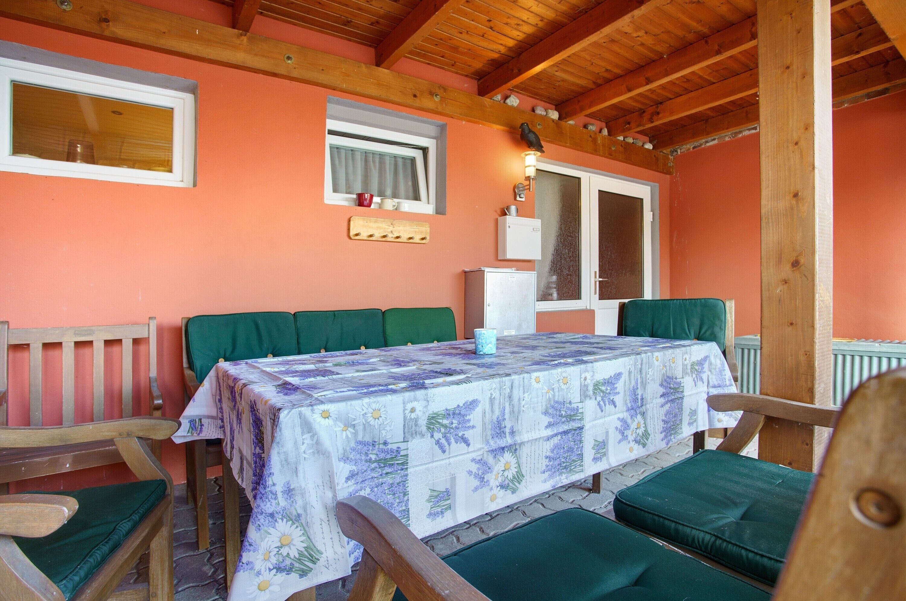 Une pièce aux murs en bois, avec une table recouverte d’une nappe à motifs floraux, des chaises rembourrées de couleur verte et une fenêtre ornée d’un rideau.