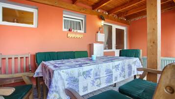 Une pièce aux murs en bois, avec une table recouverte d’une nappe à motifs floraux, des chaises rembourrées de couleur verte et une fenêtre ornée d’un rideau.