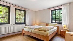 Un dormitorio con cama de madera, dos ventanas, una mesita de noche con lámpara y un radiador.