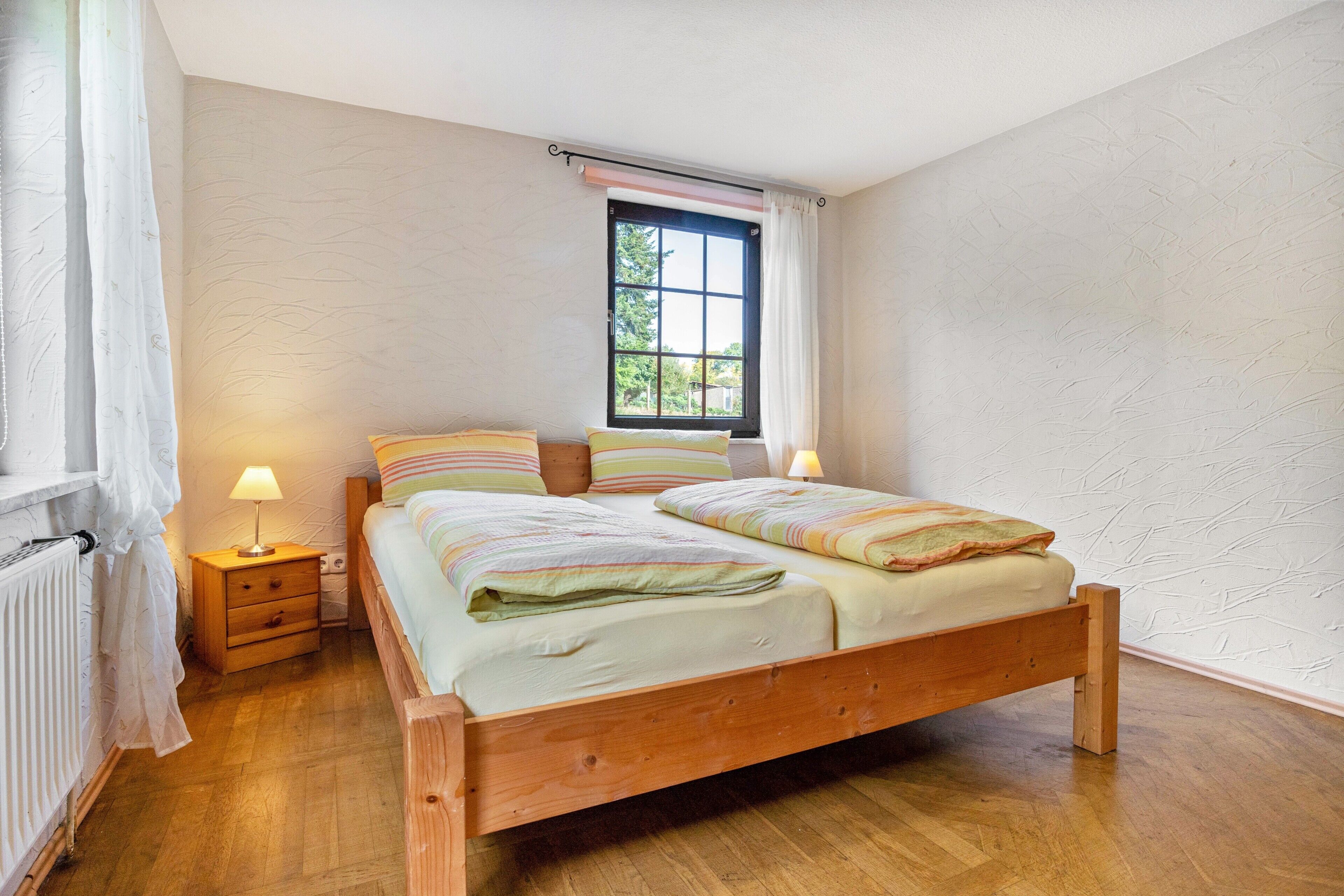 A bedroom with a wooden bed, two bedside tables, a window with curtains, and a radiator.