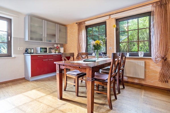 A dining area with a wooden table and chairs, a kitchen with red cabinets, and large windows with curtains.
