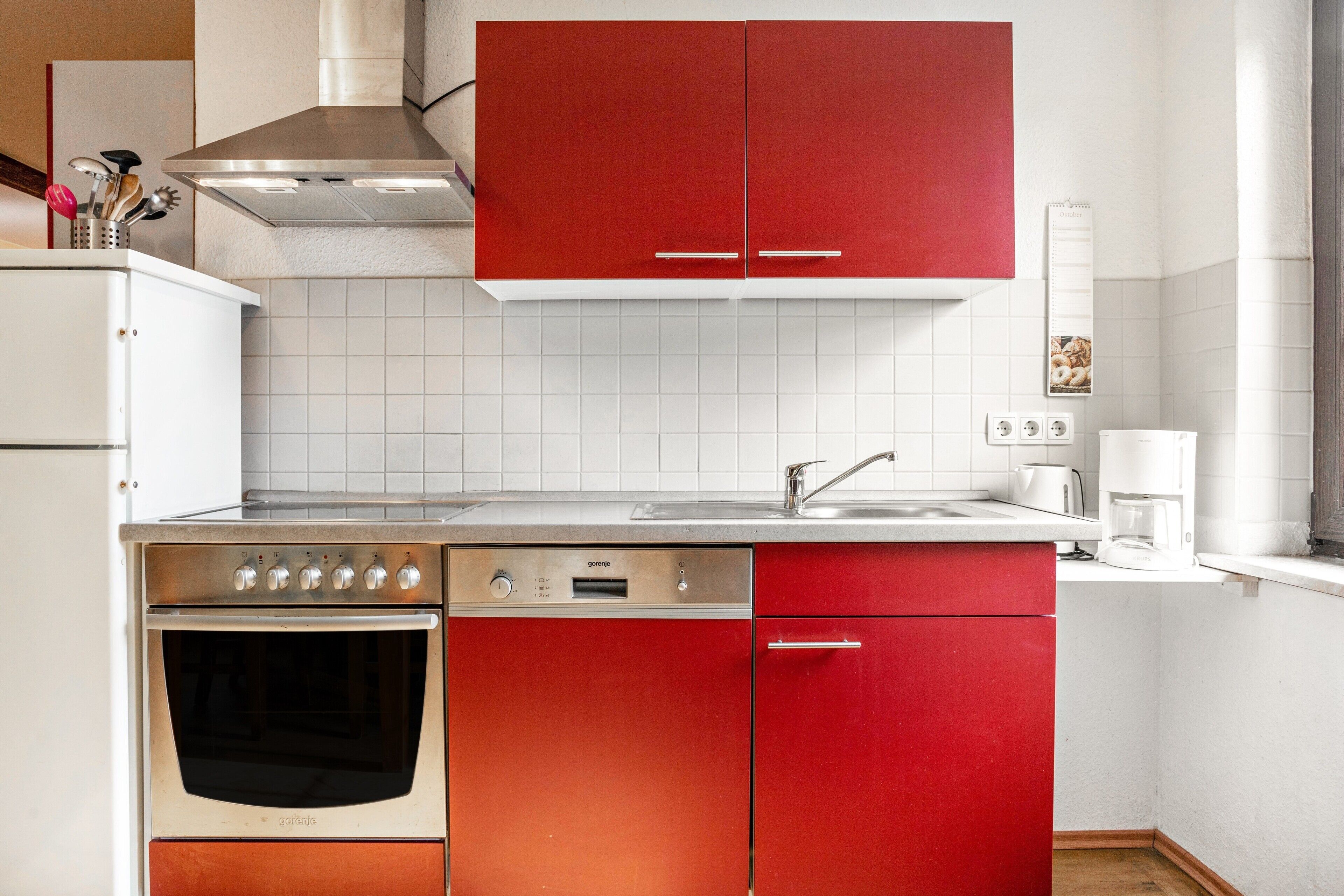 Eine Küche mit Geschirrspüler, Backofen, roten Schränken, weißen Fliesen und einer Kaffeemaschine.