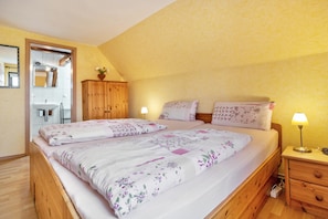 A bedroom with two beds, wooden headboards, a wooden wardrobe, a bedside table with a lamp, and a bathroom visible through an open door.