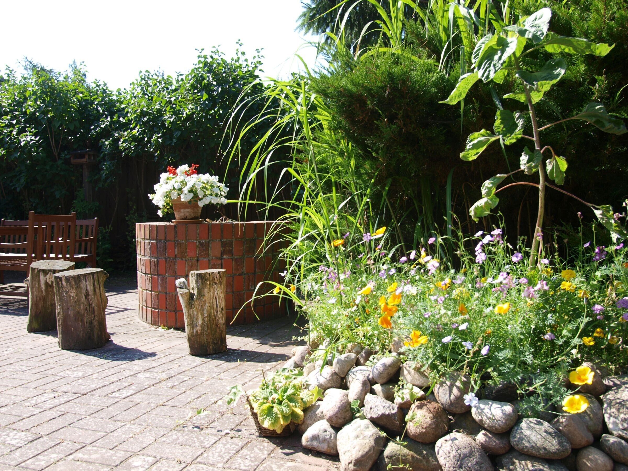 Un patio jardín con pared de ladrillo, sillas de madera y diversas plantas y flores.