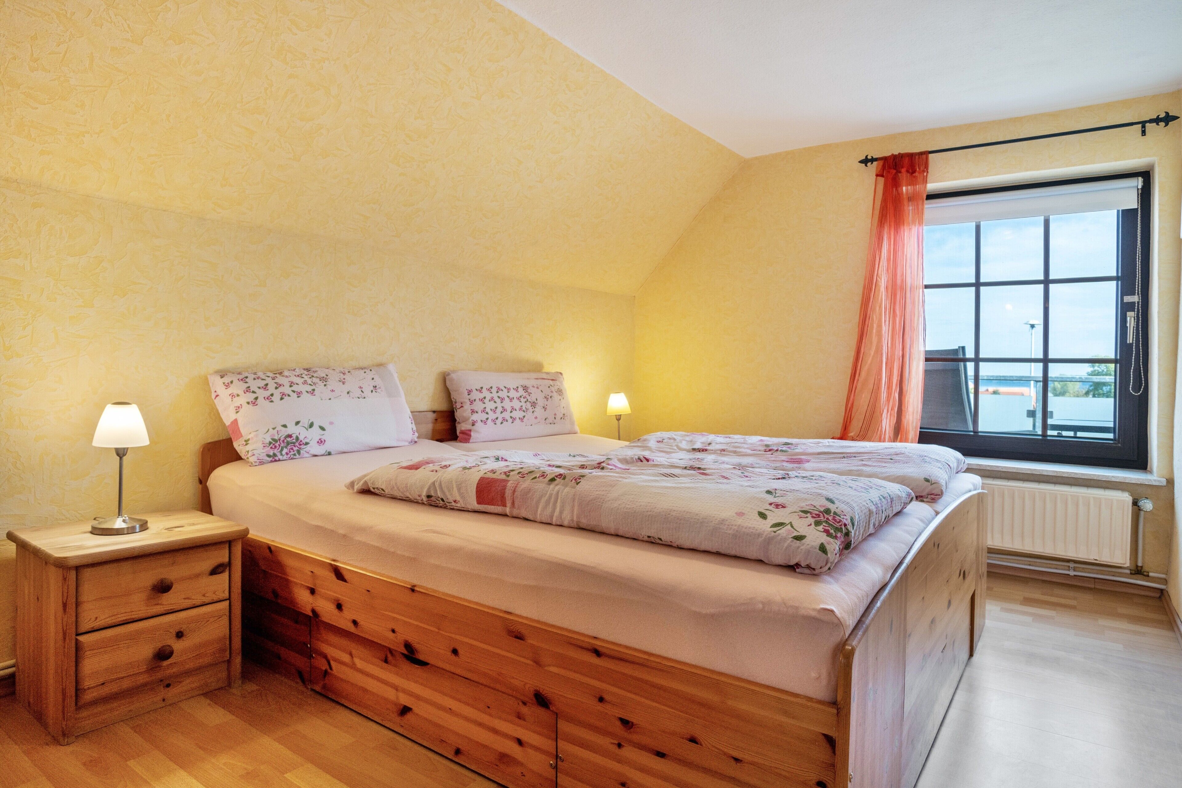 Une chambre à coucher avec un lit en bois, deux tables de chevet, une fenêtre avec des rideaux et un radiateur.