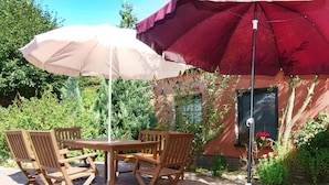 Een terras buiten met een tafel, stoelen, een grote parasol en een gebouw met een raam en planten.