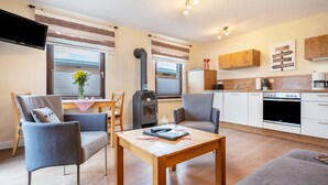 Een moderne woonkamer met een houten tafel, twee grijze fauteuils, een televisie en een keuken met witte kasten en een fornuis.